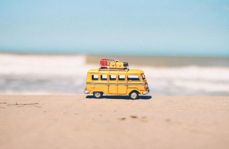 A miniature yellow toy van, a symbol of the Chevy van song era, parked on a sandy beach, representing freedom and the 1970s lifestyle.